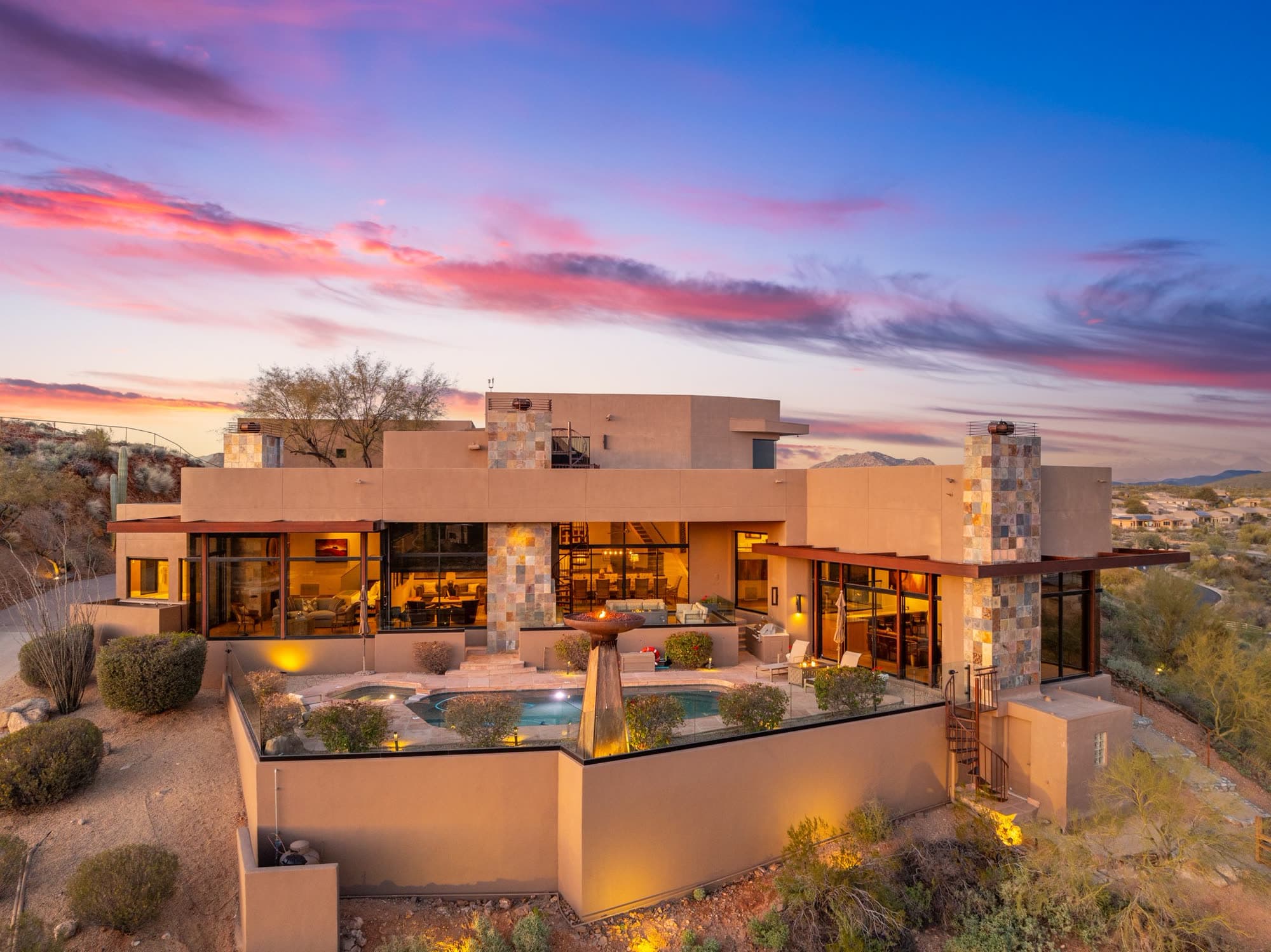 The pool, spa, and fire bowl at dusk beside the lit glass-walled interiors, under a streaked pink sky