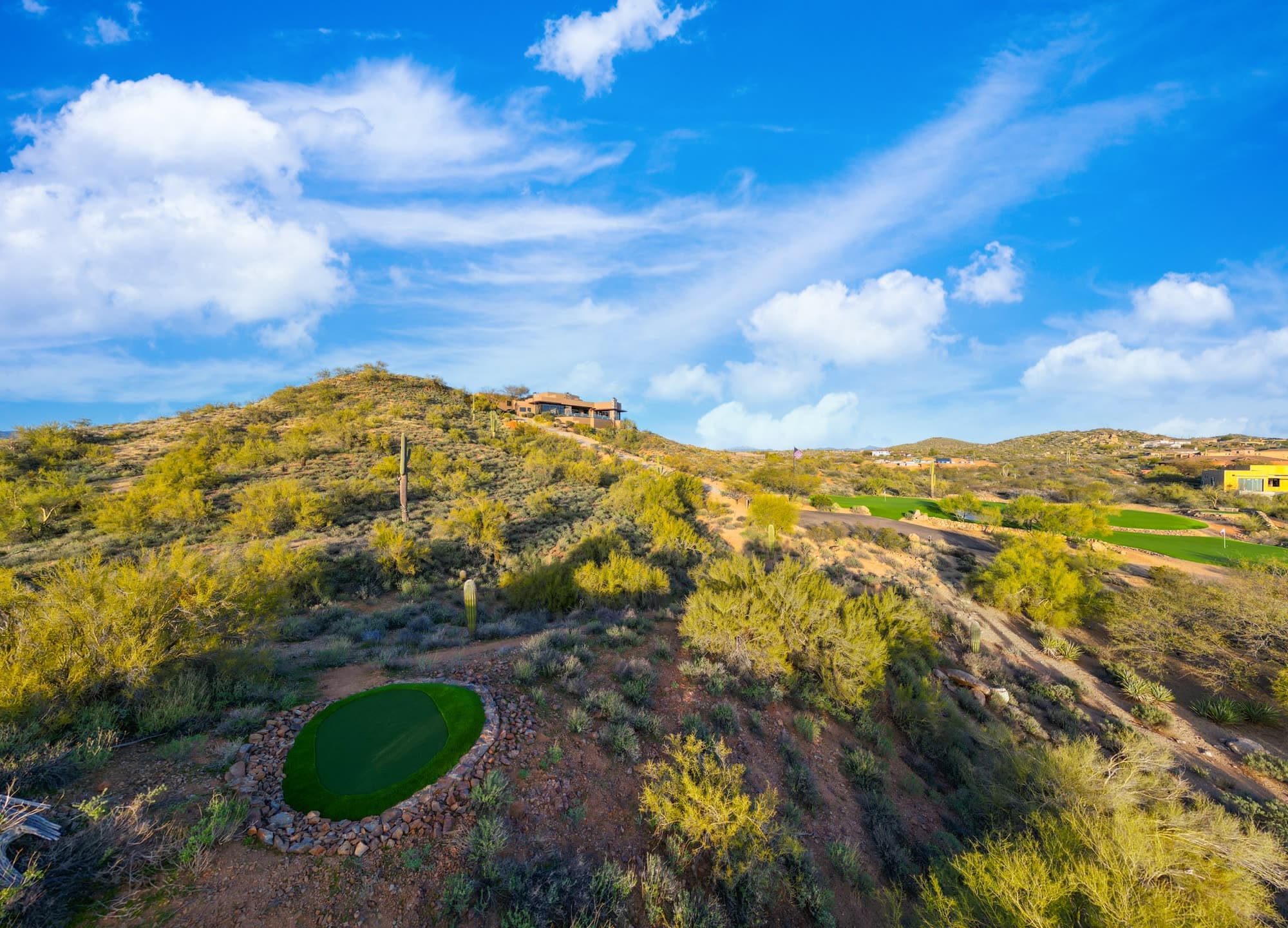 An oval satellite putting green in the foreground with the Saguaro Point house small on the ridge behind it
